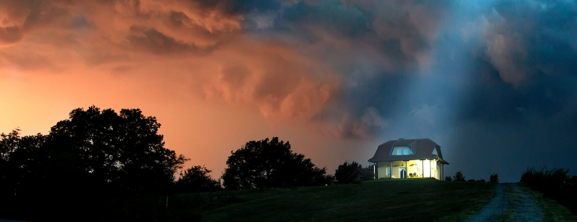 An evening storm rolls in over a house with lights on inside.